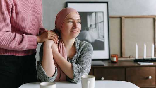 A woman in a pink headscarf sits at a table with mugs, looking thoughtful, while another person gently holds her hands in a cozy room.